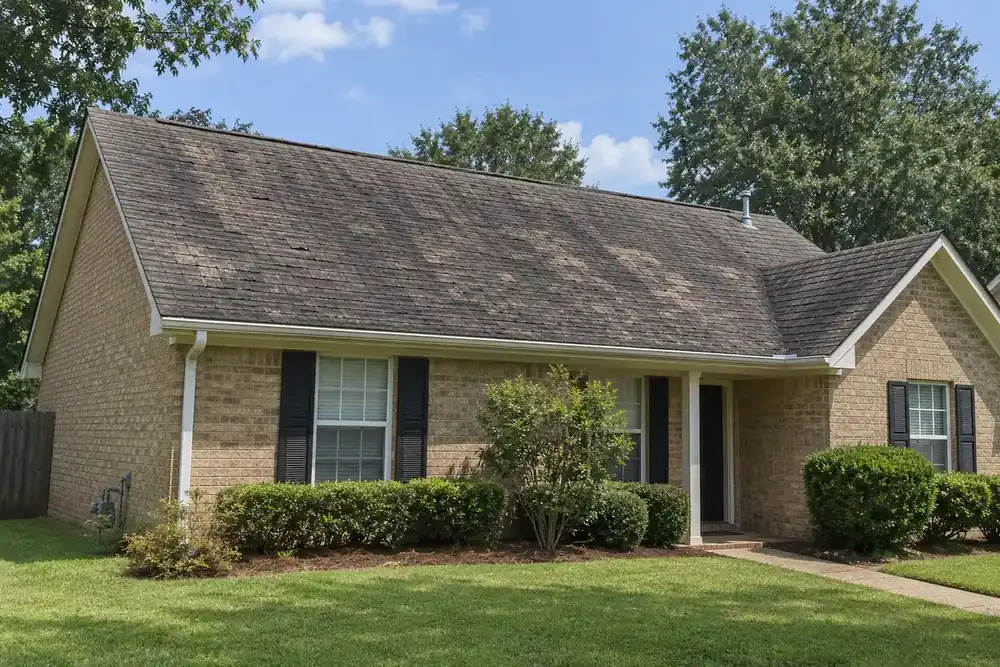 Faded, streaked and patchy shingle roof on a tan-brick ranch house with a white gutter and mown lawn